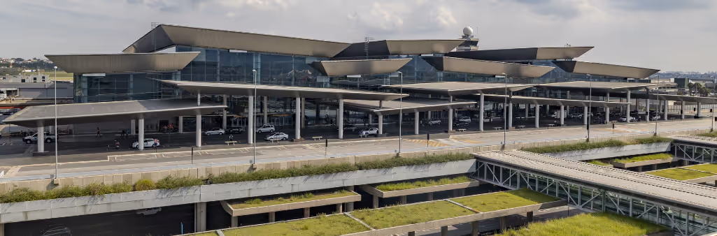 Modern airport terminal with glass facade, large canopies, and nearby parking under a cloudy sky.