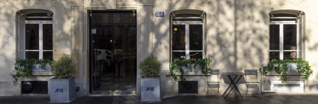 Facade of a beige building with two windows, a glass door, potted plants, and a small table with chairs.