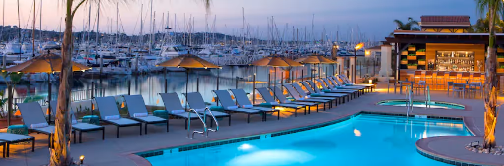Poolside lounge chairs and umbrellas by a marina with many docked boats at sunset.