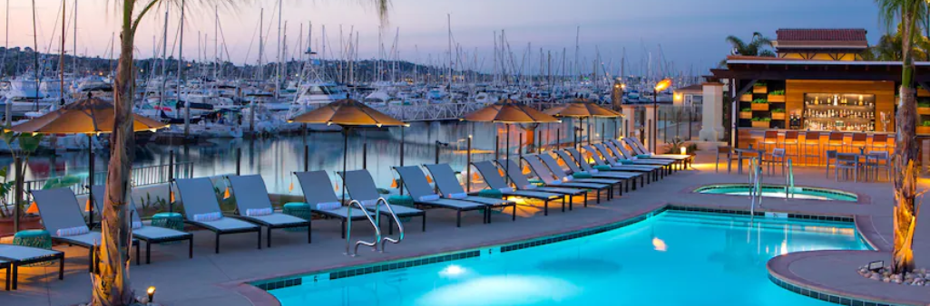 Poolside lounge chairs and umbrellas by a marina with many docked boats at sunset.