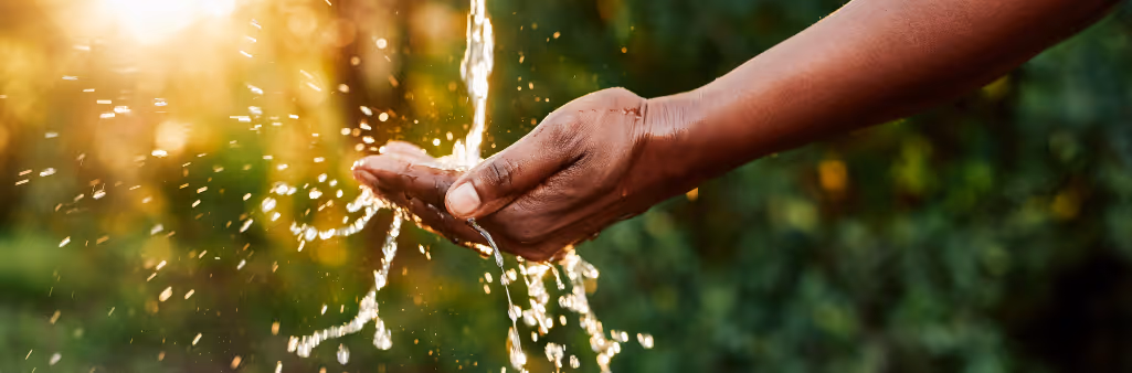 A hand catching flowing water outdoors with sunlight and greenery in the background.