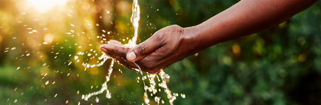 A hand catching flowing water outdoors with sunlight and greenery in the background.