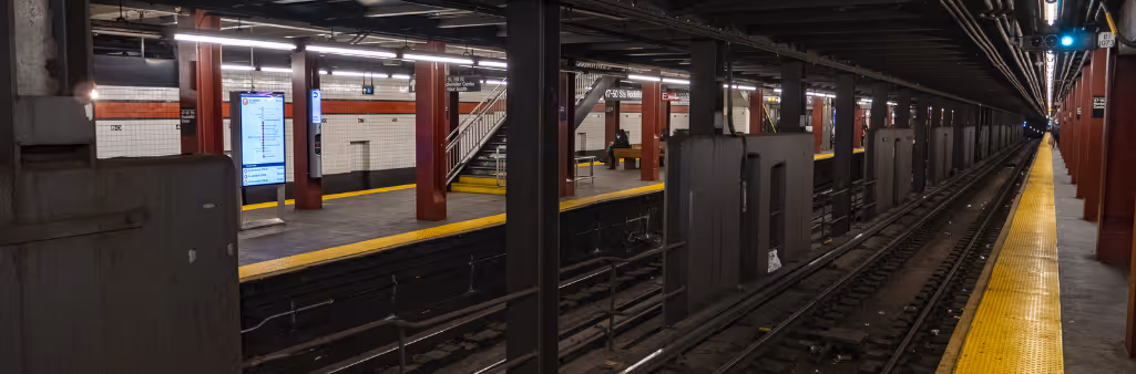 Empty subway platform with yellow safety edges, red columns, and overhead lights, viewed from track level.