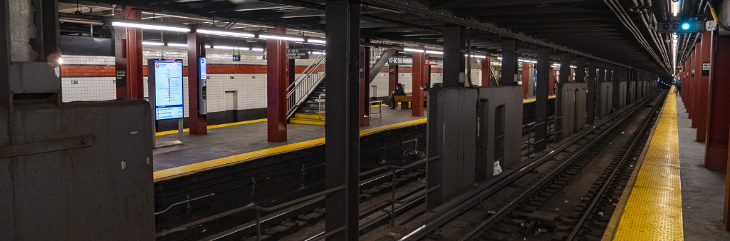 Empty subway platform with yellow safety edges, red columns, and overhead lights, viewed from track level.