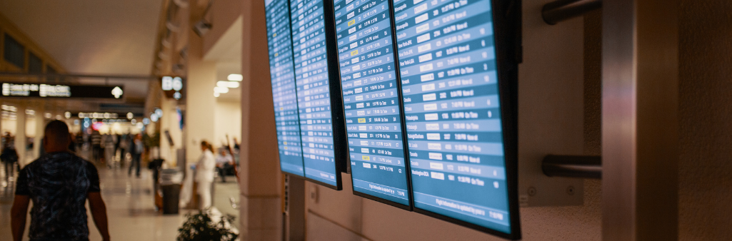 Airport hallway with people walking and several digital flight information boards mounted on the wall.