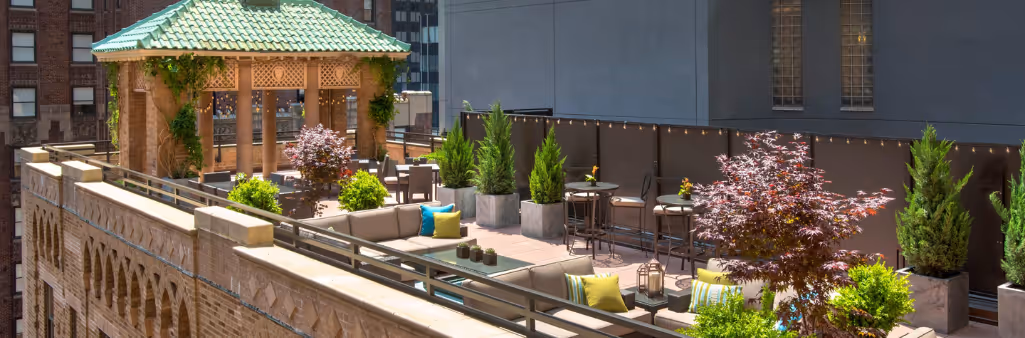 Rooftop patio with greenery, lounge seating, tables, and a gazebo, surrounded by tall city buildings.