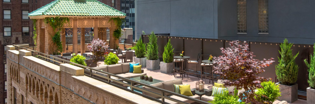 Rooftop patio with greenery, lounge seating, tables, and a gazebo, surrounded by tall city buildings.