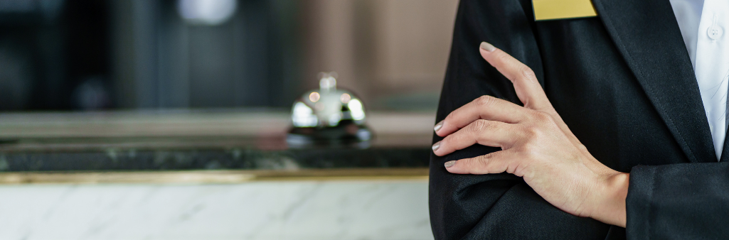 Hotel receptionist in uniform standing by a counter with a service bell in the background.