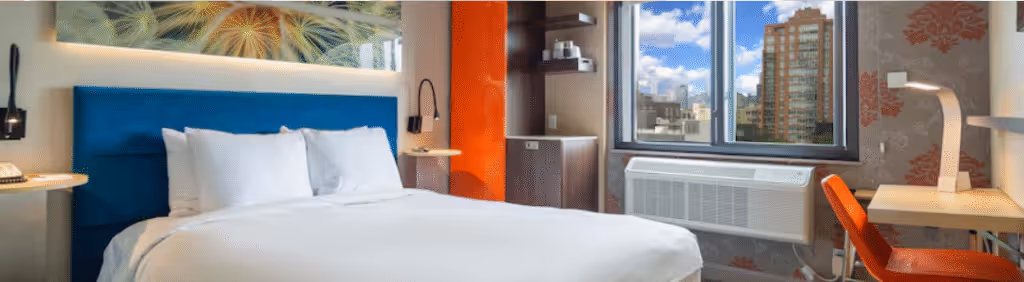 Modern hotel room with a blue headboard, white bedding, desk, orange chair, and city view through the window.