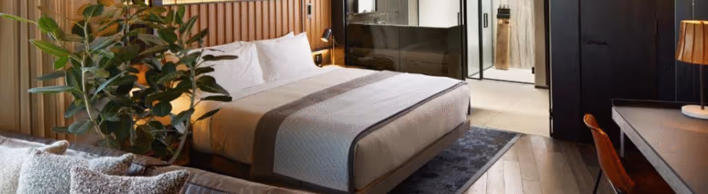 Modern hotel room with a large bed, indoor plant, desk, and a view into the bathroom area.