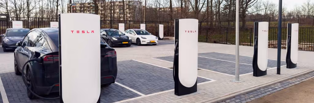 Several Tesla electric cars charging at Tesla Supercharger stations in a parking lot on a cloudy day.