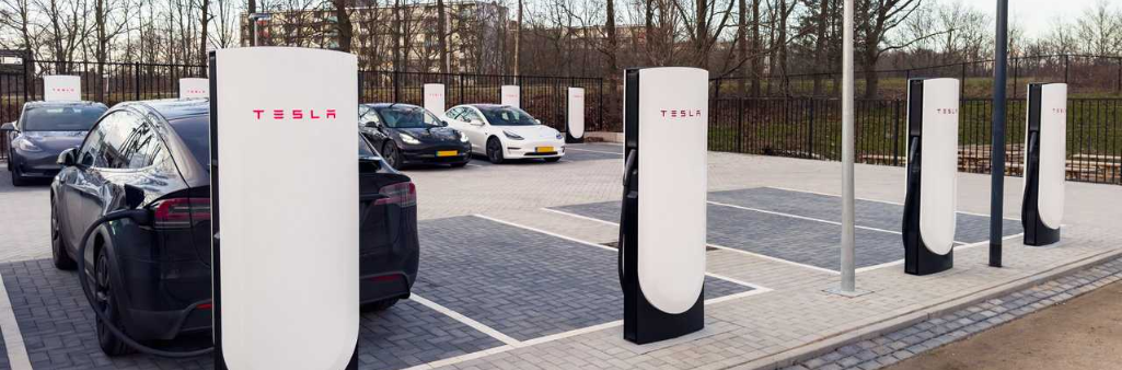 Several Tesla electric cars charging at Tesla Supercharger stations in a parking lot on a cloudy day.