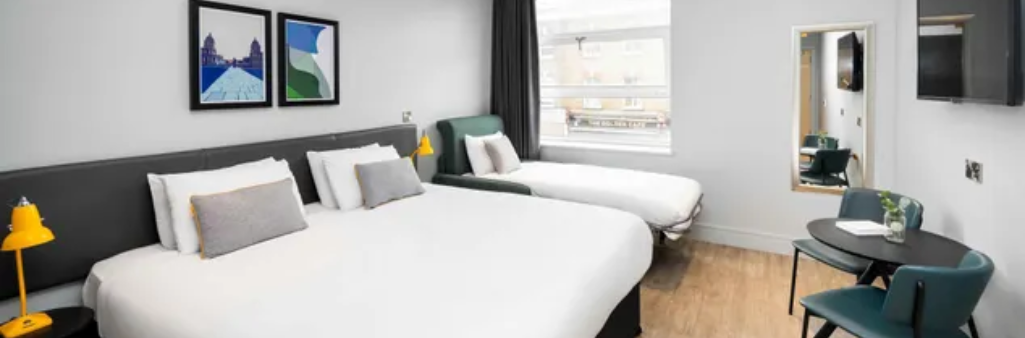 Modern hotel room with two beds, artworks, a window, a table with chairs, and a wall-mounted TV.