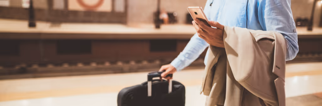 Person at a train station holding a suitcase, a coat, and a smartphone, waiting by the platform.