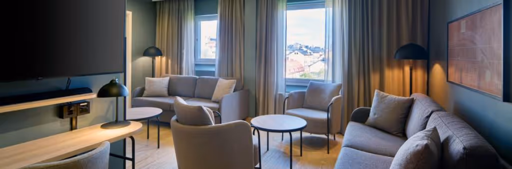 Modern living room with beige sofas, round tables, lamps, and large windows with curtains, daylight outside.
