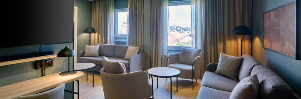 Modern living room with beige sofas, round tables, lamps, and large windows with curtains, daylight outside.