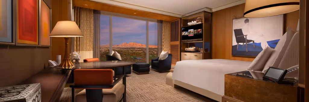 Luxurious hotel room with a king bed, seating area, desk, and large window overlooking a city and mountains at sunset.