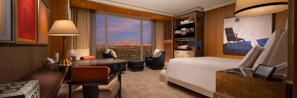 Luxurious hotel room with a king bed, seating area, desk, and large window overlooking a city and mountains at sunset.