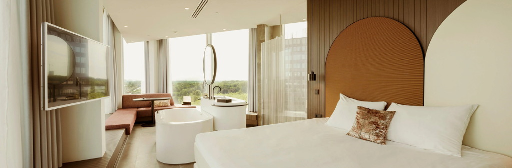 Modern hotel room with large windows, white bed, round bathtub, sofa, and city view outside.