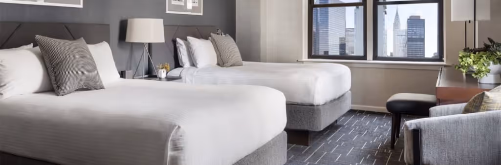 Modern hotel room with two double beds, large window, city view, desk, lamp, and neutral decor.