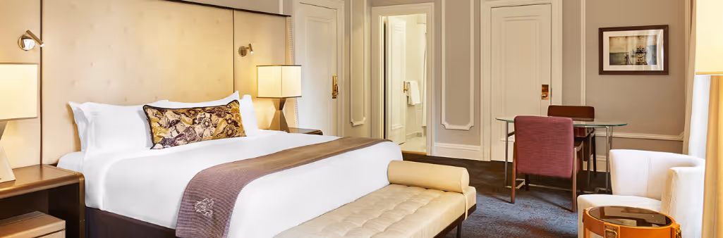 Elegant hotel room with a king bed, desk, armchair, and view into a bright bathroom.