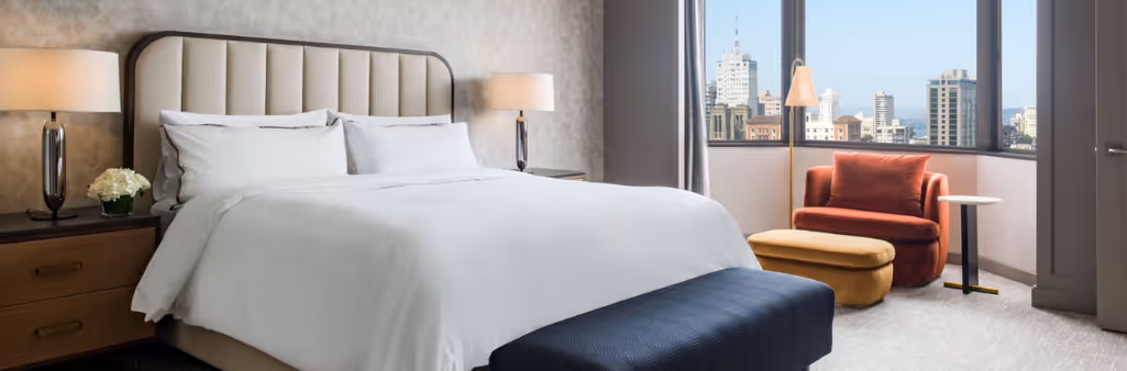 A modern hotel room with a large bed, two lamps, an orange chair, and a city view through the window.