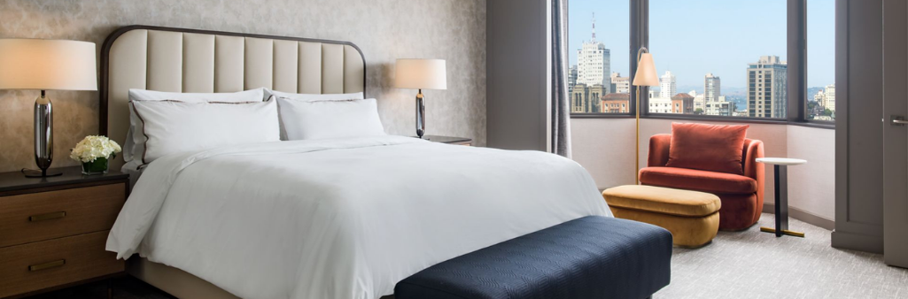 A modern hotel room with a large bed, two lamps, an orange chair, and a city view through the window.
