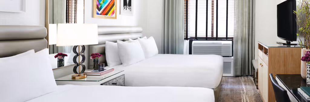 A modern hotel room with two double beds, a TV, desk, lamps, and colorful wall art above the beds.