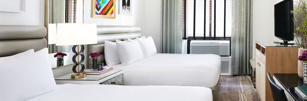 A modern hotel room with two double beds, a TV, desk, lamps, and colorful wall art above the beds.