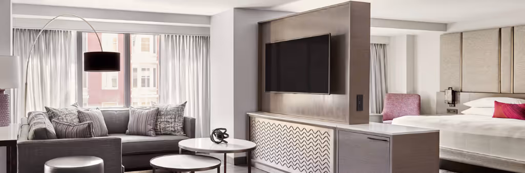 Modern hotel suite with a bed, sofa, large TV, coffee tables, and large windows with sheer curtains.