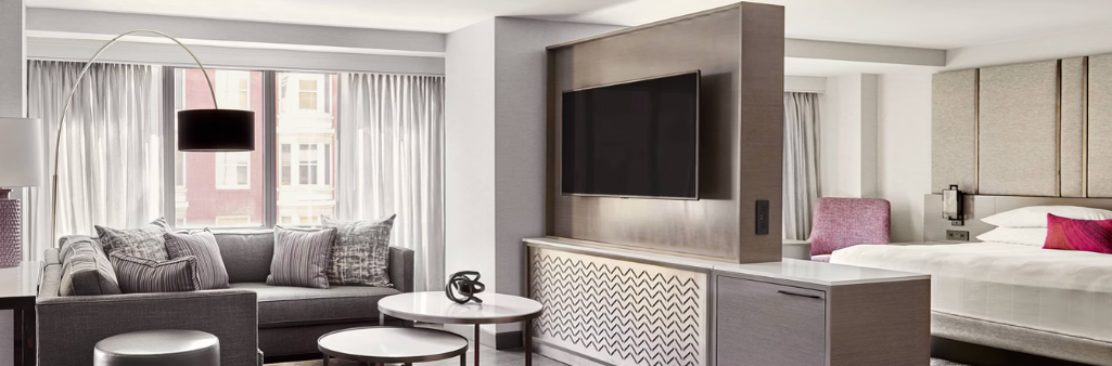 Modern hotel suite with a bed, sofa, large TV, coffee tables, and large windows with sheer curtains.