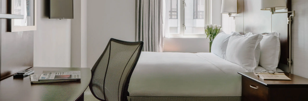 Modern hotel room with a bed, desk, chair, TV, and natural light coming through a window.