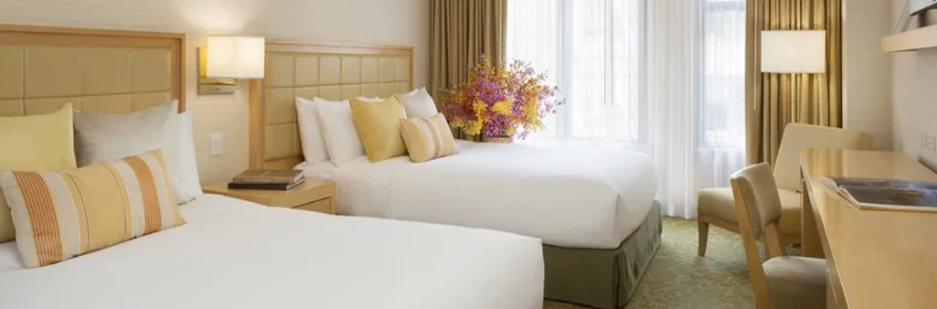 Hotel room with two double beds, light wood furniture, a desk, and a vase of flowers by the window.