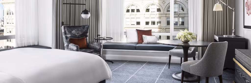 Modern hotel room with a bed, armchair, window seat, table, chairs, and large windows with city view.