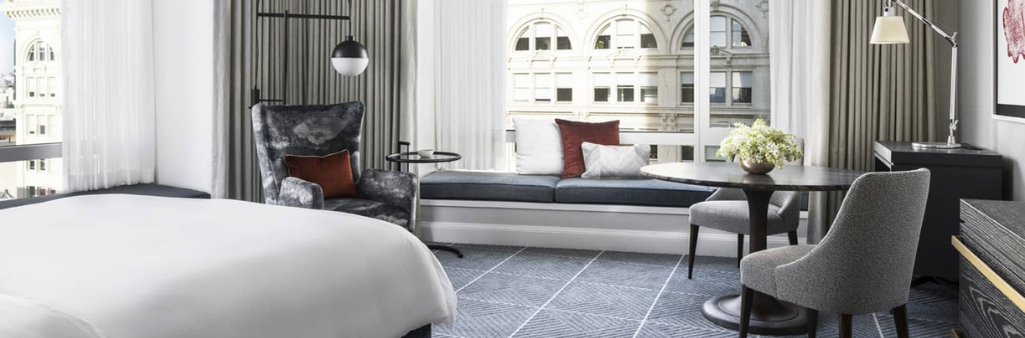 Modern hotel room with a bed, armchair, window seat, table, chairs, and large windows with city view.