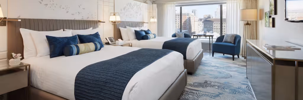 Modern hotel room with two beds, blue accents, a city view through large windows, and cozy seating area.