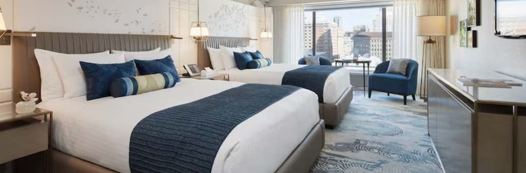 Modern hotel room with two beds, blue accents, a city view through large windows, and cozy seating area.