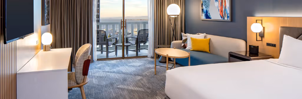 Modern hotel room with a bed, sofa, desk, balcony, and city view through sliding glass doors.