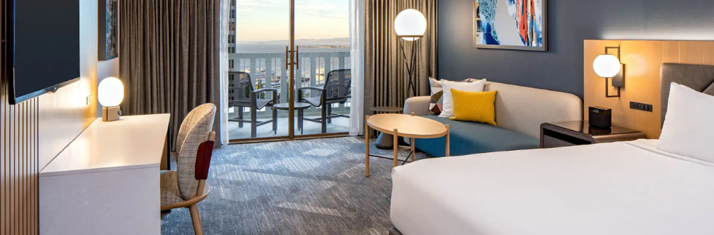 Modern hotel room with a bed, sofa, desk, balcony, and city view through sliding glass doors.