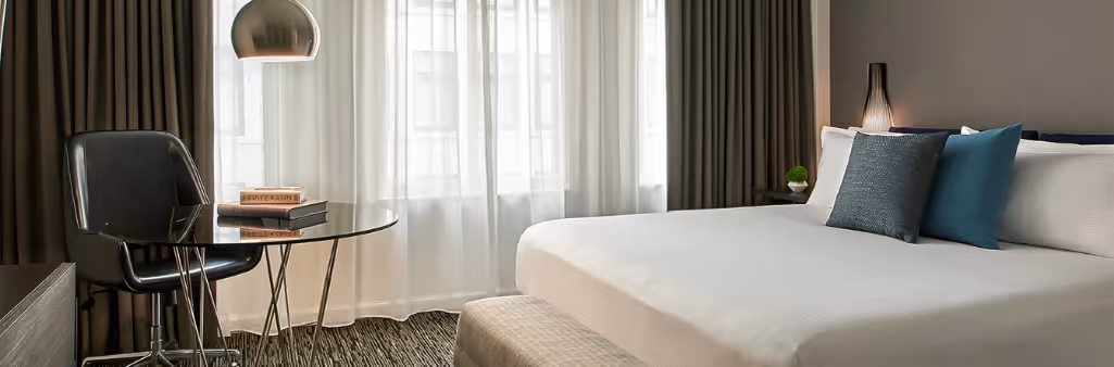 Modern hotel room with a large bed, glass desk, black chair, and window with curtains letting in natural light.