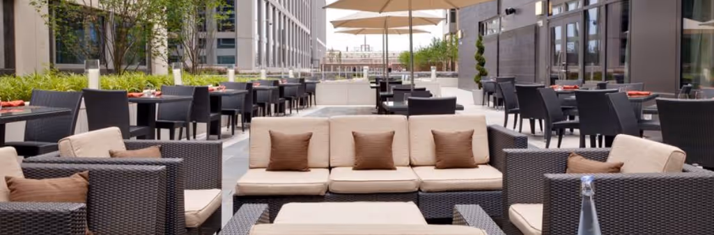 Outdoor patio with wicker sofas, dining tables, umbrellas, and greenery in a modern urban setting.