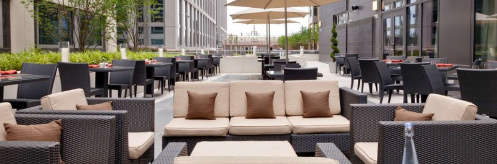 Outdoor patio with wicker sofas, dining tables, umbrellas, and greenery in a modern urban setting.