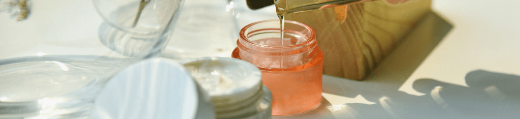 A close-up of a clear liquid being poured into a small, open, pink container with an Honest Company gift card nearby. Other empty jars and a wooden box rest on the sunlit surface.