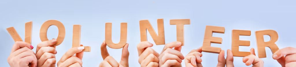 Multiple hands holding up wooden letters spelling VOLUNTEER against a light blue background.