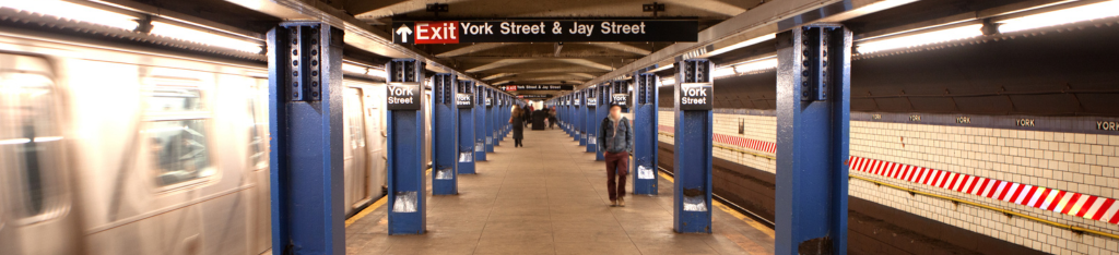 Blurred subway train departing York Street station, blue columns, tiled walls, and people on the platform.