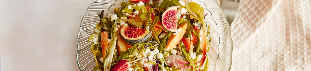 A glass plate of salad with figs, greens, carrots, and crumbled cheese on a light tablecloth.