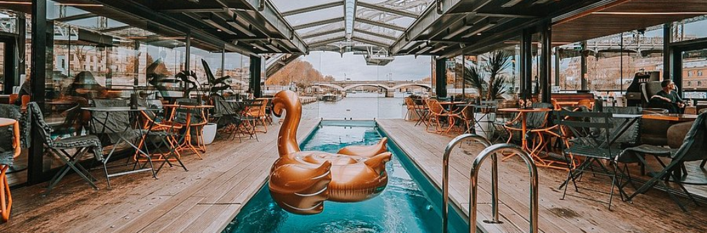 Open-air café with a central pool, swan float, and river view through large windows on both sides.