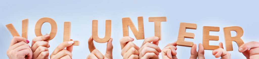 Multiple hands holding up wooden letters spelling VOLUNTEER against a light blue background.