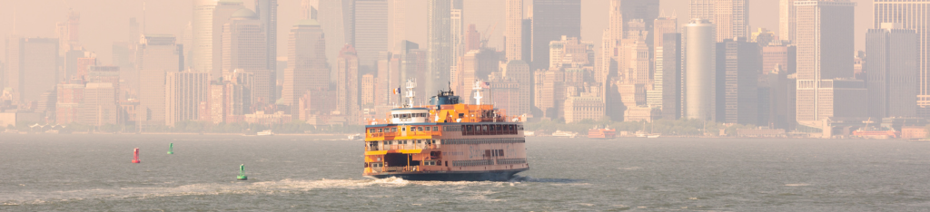An orange Staten Island Ferry sails toward Manhattan with a hazy city skyline in the background.