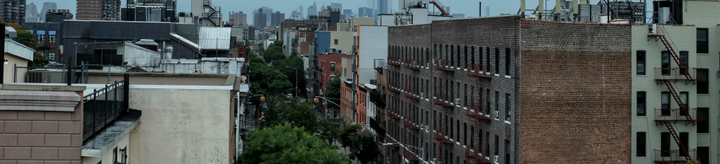 View of a city street lined with apartment buildings, trees, and skyscrapers in the distance under a cloudy sky.